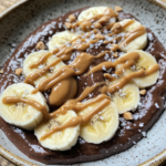 Chocolate protein pudding topped with banana slices, peanut butter drizzle, and crushed nuts.