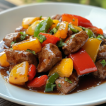 Close-up of a beef stir-fry with colorful bell peppers, coated in a rich, glossy sauce and garnished with fresh herbs, served on a white plate.